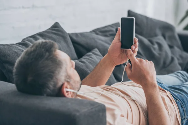 Взрослый мужчина слушает музыку в наушниках и держит смартфон с пустым экраном, лежа на диване — стоковое фото