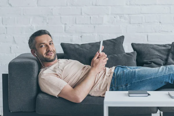 Handsome man smiling at camera while lying on sofa, listening music in earphones and holding smartphone — Stock Photo