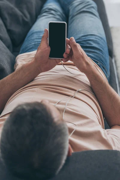 Selective focus of adult man listening music in earphones and holding smartphone with blank screen while lying on sofa — Stock Photo