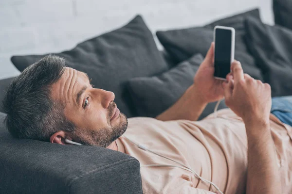 Smiling man in earphones lying on sofa and holding smartphone with blank screen — Stock Photo