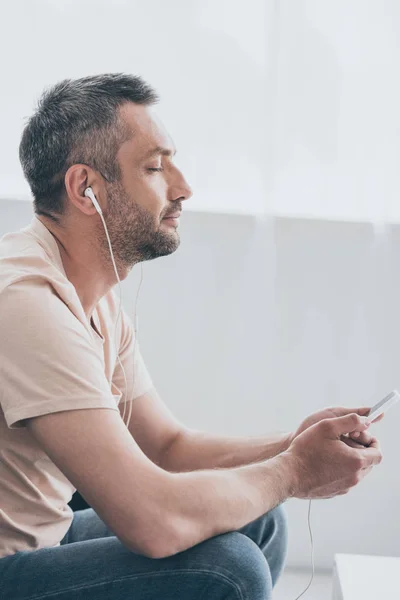 Schöner Mann mit geschlossenen Augen, der in Kopfhörern Musik hört, während er sein Smartphone hält — Stockfoto