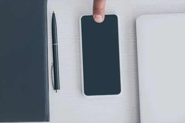 Обрезанный вид мужского пальца на смартфоне с чистым экраном рядом с ноутбуком, ноутбуком и ручкой — стоковое фото