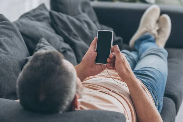 Selective focus of man in earphones lying on sofa and holding smartphone with blank screen — Stock Photo