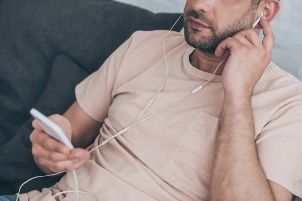 Обрезанный вид взрослого мужчины, держащего смартфон и слушающего музыку в наушниках — стоковое фото