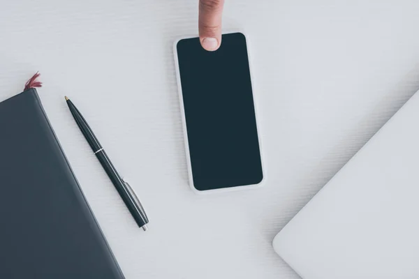 Visão parcial do dedo masculino no smartphone com tela em branco perto do notebook, laptop e caneta — Fotografia de Stock