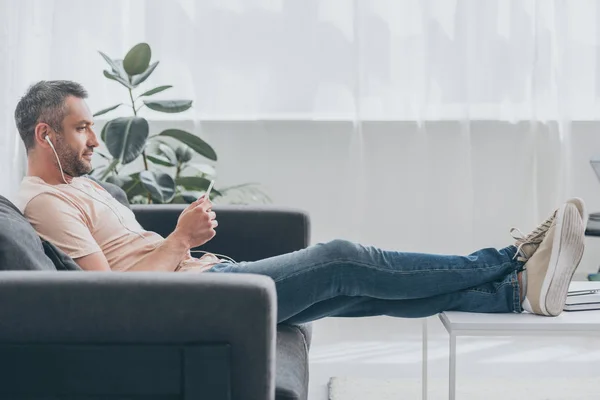 Handsome man in earphones using smartphone while sitting on sofa with legs on table — Stock Photo