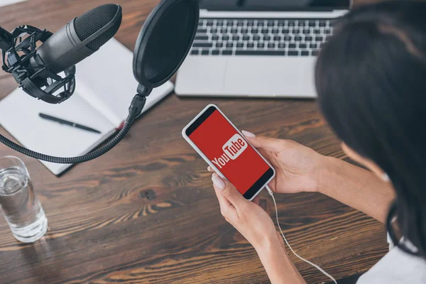 KYIV, UCRAINA - 5 MAGGIO 2019: Vista parziale o donna che tiene smartphone con app Youtube sullo schermo vicino a microfono e laptop . — Foto stock