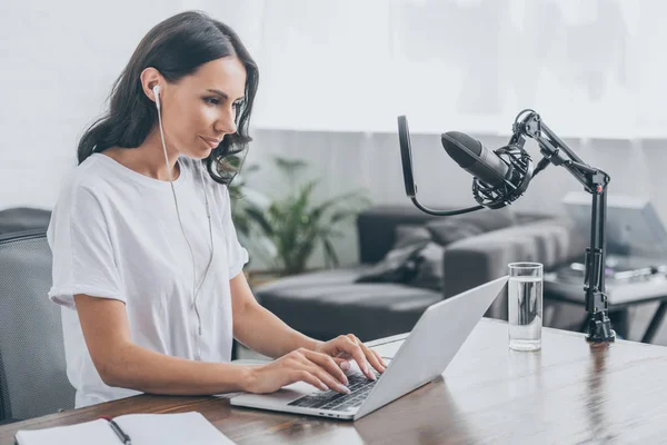 Pretty radio host in earphones using laptop while sitting at workplace near microphone — Stock Photo