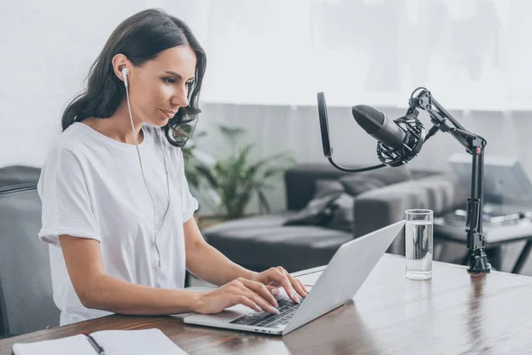 Anfitrión de radio bonita en los auriculares usando el ordenador portátil mientras está sentado en el lugar de trabajo cerca del micrófono - foto de stock