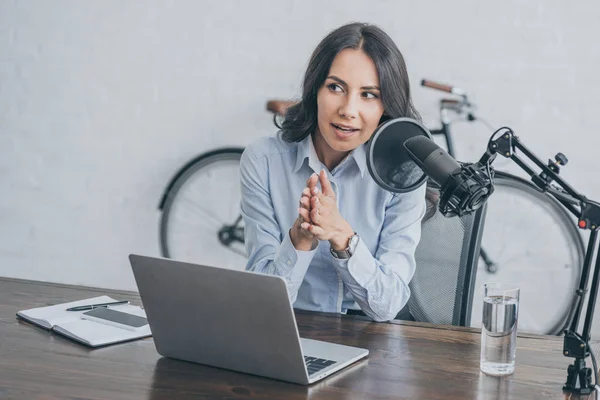 Hübsche Radiomoderatorin spricht in Mikrofon, während sie am Schreibtisch neben dem Laptop sitzt — Stockfoto