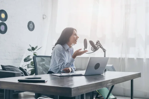 Junger Radiomoderator nimmt Podcast auf, während er am Holztisch neben Mikrofon und Laptop sitzt — Stockfoto