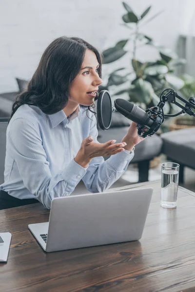 Привлекательный радиоведущий, говорящий в микрофоне, сидя за деревянным столом рядом с ноутбуком — стоковое фото