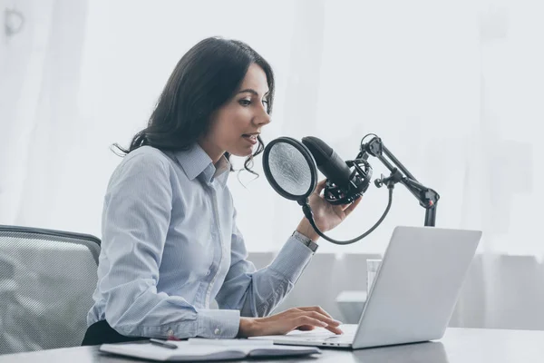 Anfitrión de radio bonita usando el ordenador portátil mientras habla en micrófono en el lugar de trabajo en estudio de radiodifusión - foto de stock