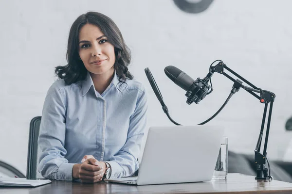 Bella radio host guardando la fotocamera mentre seduto sul posto di lavoro vicino a microfono e laptop — Foto stock