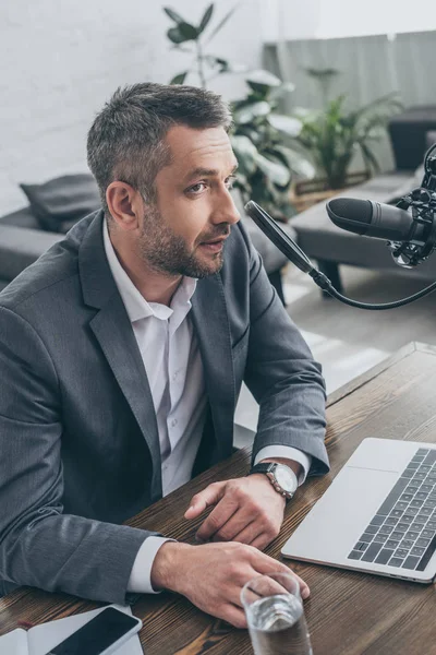 Schöner Radiomoderator spricht in Mikrofon, während er am Arbeitsplatz in der Nähe von Laptop sitzt — Stockfoto