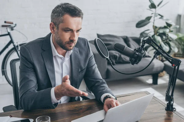 Beau hôte radio en utilisant un ordinateur portable tout en étant assis sur le lieu de travail près du microphone — Photo de stock