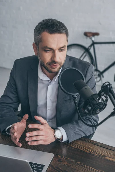 Serio gesto de anfitrión de radio mientras habla en micrófono en el lugar de trabajo cerca de la computadora portátil - foto de stock