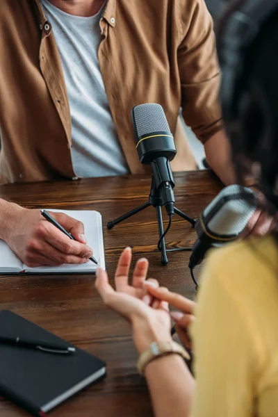 Vista recortada de la escritura del anfitrión de radio en el cuaderno mientras entrevista a la mujer en el estudio de radiodifusión - foto de stock