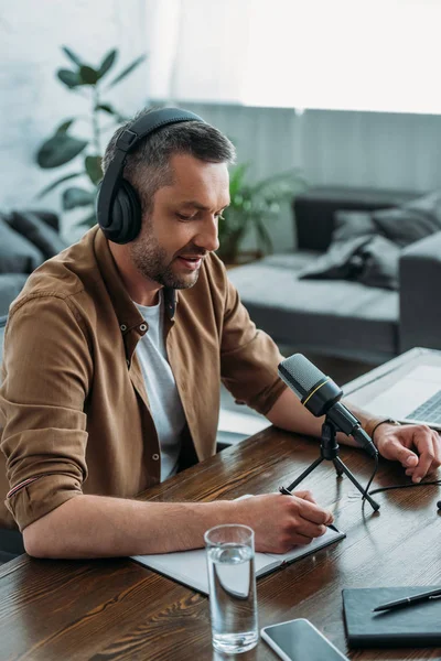 Anfitrião de rádio bonito em fones de ouvido escrevendo em notebook enquanto sentado no local de trabalho perto de microfone — Fotografia de Stock