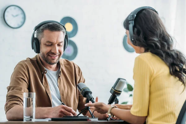 Cheerful radio host in headphones writing in notebook while sitting at workplace near colleague — Stock Photo