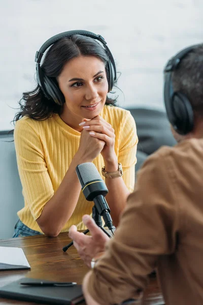 Избирательный фокус внимания радиоведущего в наушниках, смотрящего на коллегу, сидящего рядом с микрофоном в студии вещания — стоковое фото