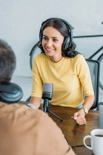 Foco seletivo de anfitrião de rádio sorridente em fones de ouvido olhando para colega enquanto sentado perto de microfone no estúdio de radiodifusão — Fotografia de Stock