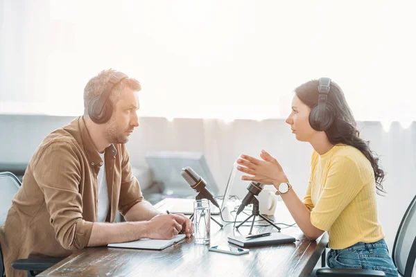 Zwei seriöse Radiomoderatoren unterhalten sich während sie im Sendestudio neben Mikrofonen sitzen — Stockfoto