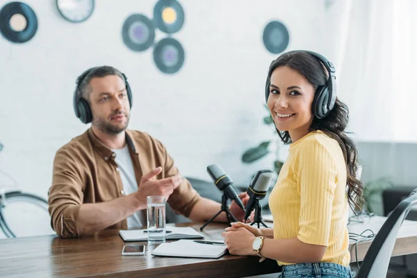 Anfitrión de radio alegre mirando a la cámara mientras está sentado en el lugar de trabajo cerca de colega - foto de stock