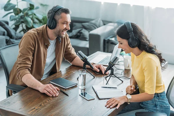 Zwei lächelnde Radiomoderatoren mit Kopfhörern im Gespräch, während sie am Arbeitsplatz in der Nähe von Mikrofonen sitzen — Stockfoto