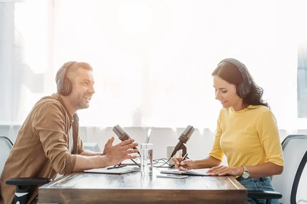 Two smiling radio hosts in headphones talking while sitting at table in studio — Stock Photo