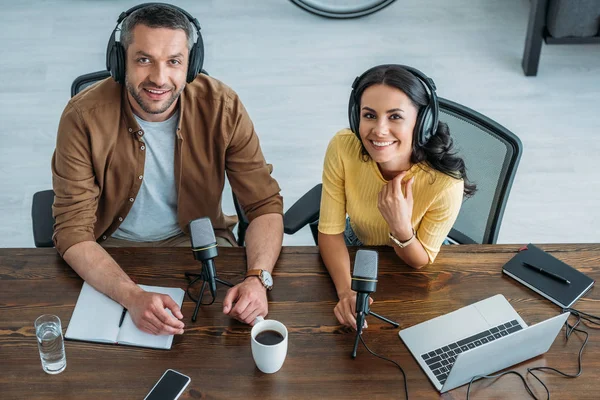 Two cheerful radio hosts in headphones sitting at workplace and smiling at camera — Stock Photo