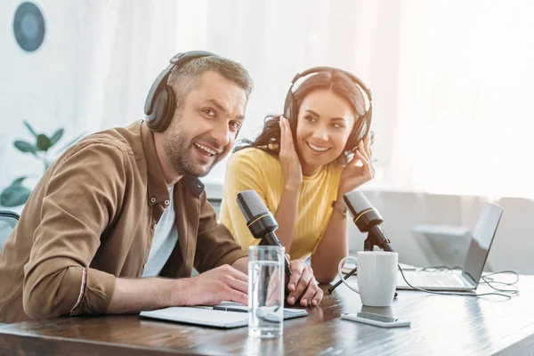 Anfitrião de rádio alegre olhando para a câmera enquanto sentado perto de colega bonito — Fotografia de Stock