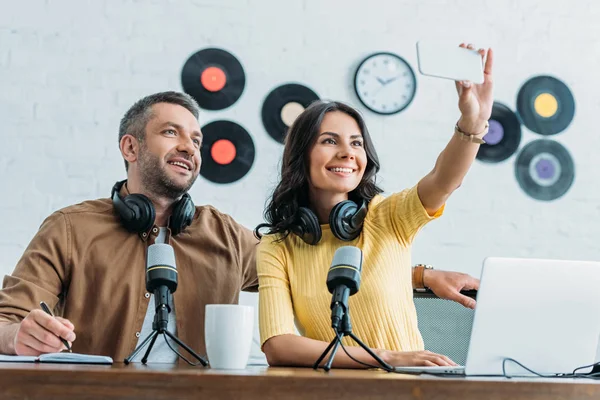 Atractivo anfitrión de radio sentado cerca de colega guapo y tomar selfie con teléfono inteligente - foto de stock