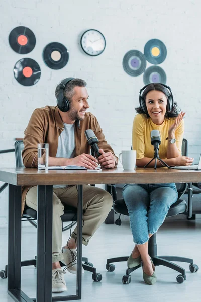 Dos anfitriones de radio sonrientes sentados en el lugar de trabajo en el estudio de radiodifusión y grabación podcast juntos - foto de stock