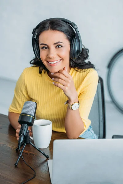 Jolie animateur radio dans les écouteurs souriant à la caméra tout en étant assis près du microphone — Photo de stock