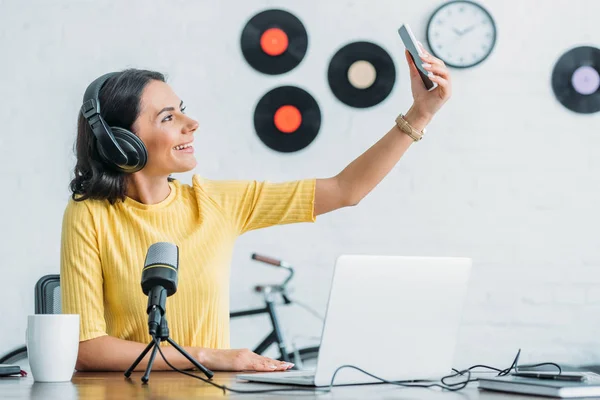 Attraktiver Radiomoderator mit Kopfhörer, der ein Selfie mit dem Smartphone macht, während er neben dem Mikrofon sitzt — Stockfoto