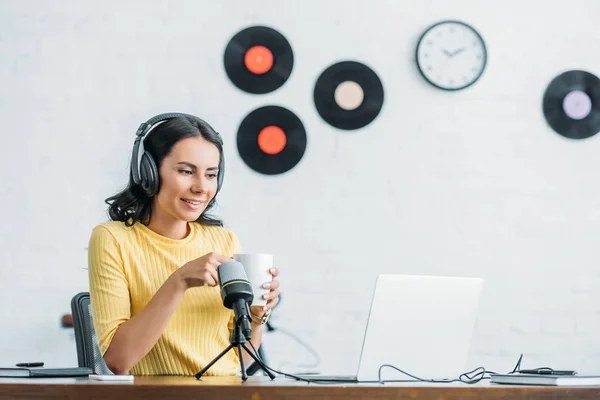 Pretty radio host in headphones looking at laptop while holding coffee cup — Stock Photo
