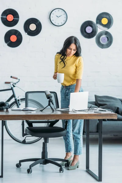 Pretty radio host using laptop and holding coffee cup while standing at wokplace in studio — Stock Photo