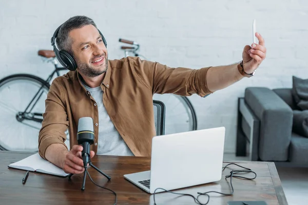 Anfitrião de rádio bonito em fones de ouvido tirar selfie com smartphone enquanto sentado no local de trabalho — Fotografia de Stock