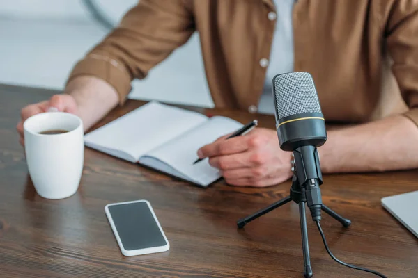 Vista ritagliata di scrittura host radio in notebook mentre seduto alla scrivania vicino smartphone con schermo bianco e tazza di caffè — Foto stock