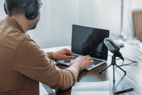 Радиоведущий в наушниках, используя ноутбук с экраном, сидя рядом с микрофоном — стоковое фото
