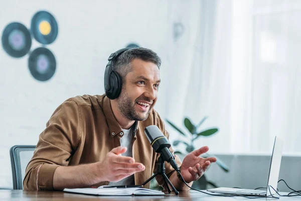 Anfitrião de rádio sorridente em fones de ouvido gesticulando enquanto fala em microfone no estúdio de radiodifusão — Fotografia de Stock