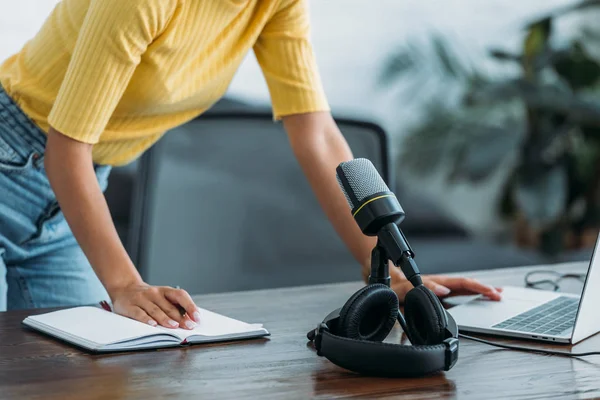 Partial view of radio host standing at workplace near microphone, laptop, headphones and notebook — Stock Photo