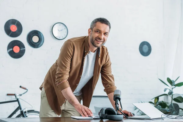 Schöner Radiomoderator lächelt in die Kamera, während er am Arbeitsplatz im Sendestudio steht — Stockfoto