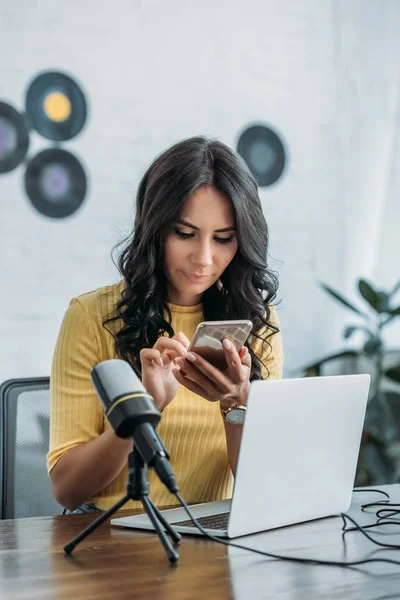 Beautiful radio host using smartphone while sitting near microphone and laptop — Stock Photo