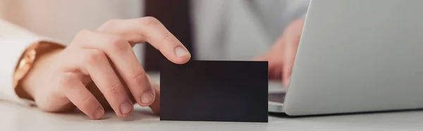 Partial view of businessman holding empty black business card while using laptop in office, panoramic shot — Stock Photo