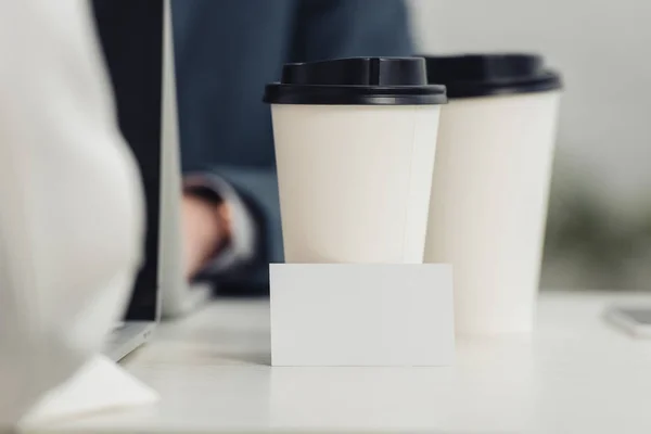 Selective focus of blank business card and disposable cups on table near businesspeople using laptops — Stock Photo