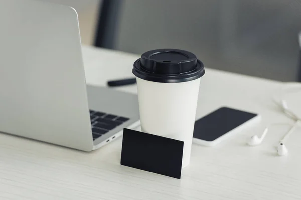 Cartão de visita preto vazio, copo de papel, laptop e smartphone com tela em branco na mesa de escritório — Fotografia de Stock