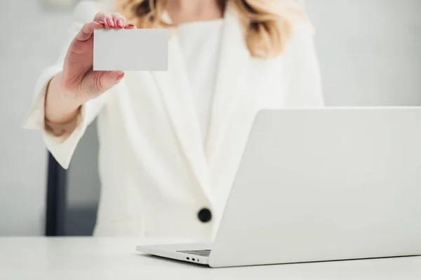 Vista recortada de la mujer de negocios que muestra en la cámara blanca tarjeta de visita en blanco mientras está sentado cerca de la computadora portátil - foto de stock