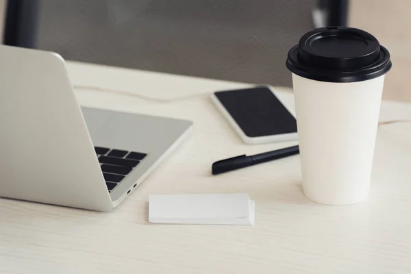 Tarjetas de visita vacías blancas, ordenador portátil, café para llevar, pluma y teléfono inteligente con pantalla en blanco en la mesa de la oficina - foto de stock